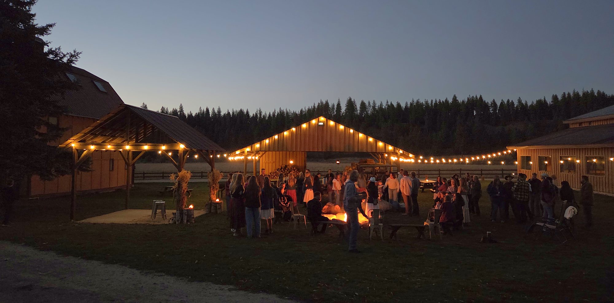 Guests around the fire at dusk on October 3, string lights on the wood shed and The Round Table